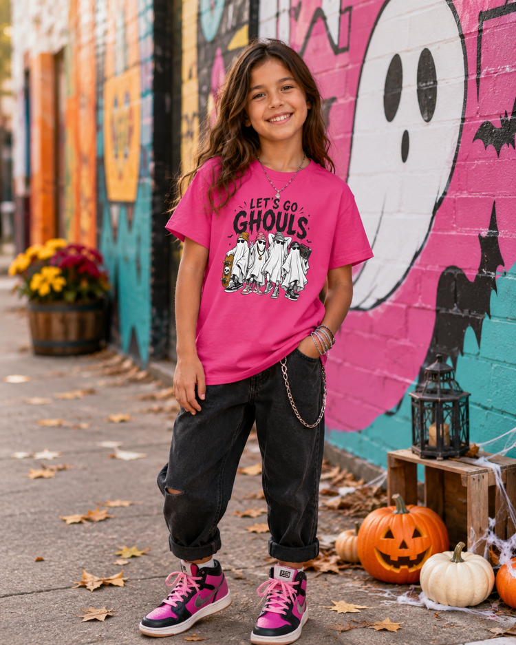 A young girl stands outdoors in front of a colorful Halloween-themed mural, wearing a bright pink graphic T-shirt that reads 'LET'S GO GHOULS' with a ghost and skeleton design, dark ripped jeans, and pink-and-black sneakers.