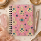 A pink spiral notebook with a Hamsa hand and eye pattern, styled in a boho flat-lay with crystals, dried flowers, a candle, and a pen.