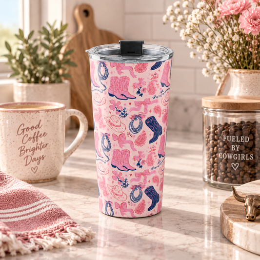 A pink insulated travel tumbler with a cowgirl-themed pattern of bows, boots, and horseshoes, standing on a marble kitchen counter surrounded by lifestyle props like a coffee mug, a jar of coffee beans, and flowers.