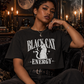 A woman sits on a dark velvet armchair in a dimly lit, gothic room, wearing a black graphic t-shirt with 'BLACK CAT ENERGY' and a white cat illustration printed on the front.