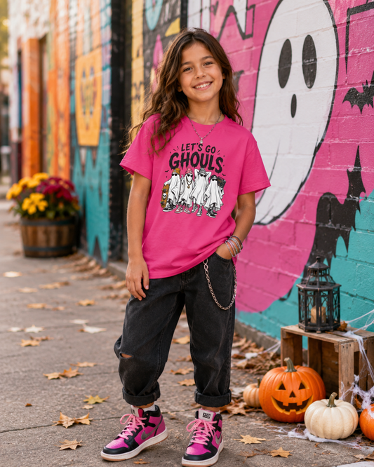 A young girl stands outdoors in front of a colorful Halloween-themed mural, wearing a bright pink graphic T-shirt that reads 'LET'S GO GHOULS' with a ghost and skeleton design, dark ripped jeans, and pink-and-black sneakers.